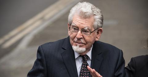 Rolf Harris in a suit and glasses waving.