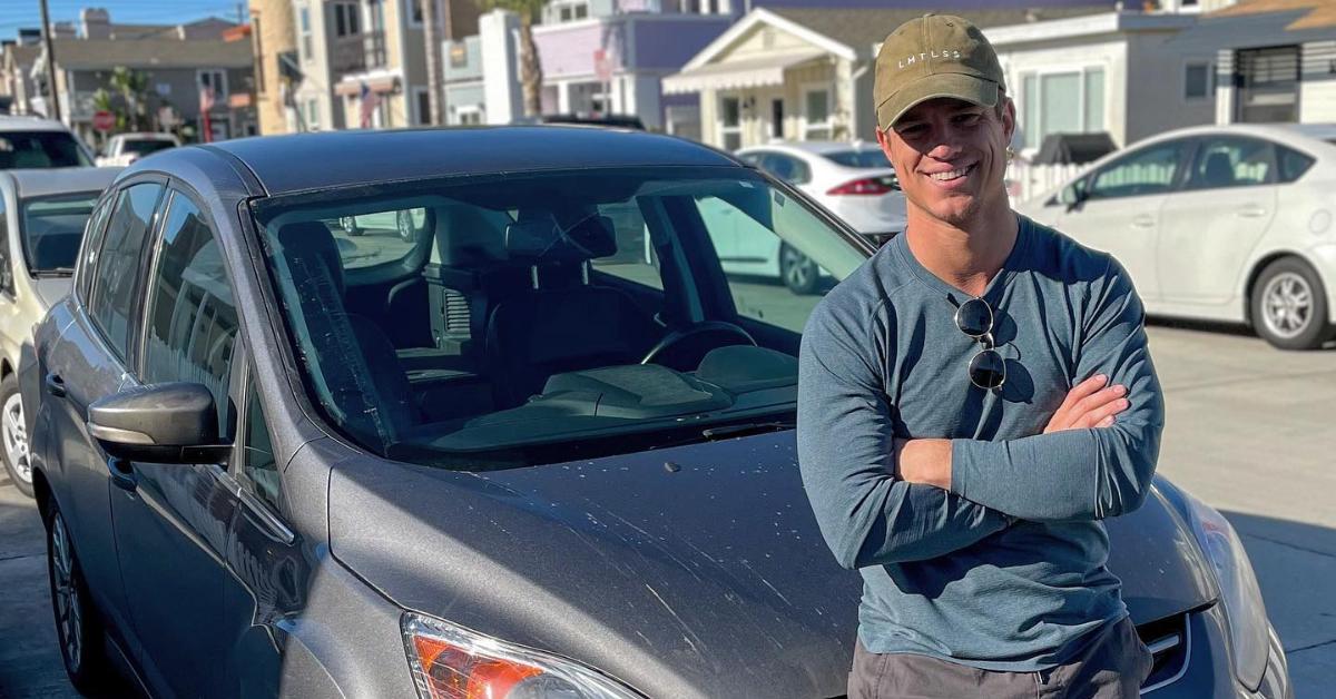Ben Mezzenga, a rumored contestant for Season 7 of 'Love Is Blind,' stands casually leaning against his car, arms crossed, wearing gray pants, a blue long-sleeve shirt, and a green baseball hat, all while flashing a smile.