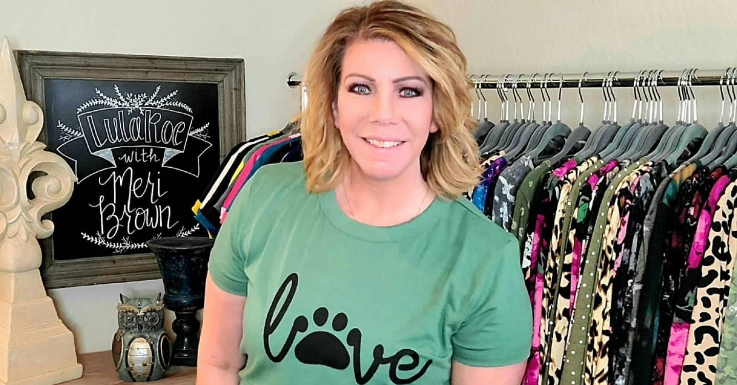 Meri Brown smiles and poses next to a rack of clothes wearing a green shirt with a pawprint that says "love" in black.