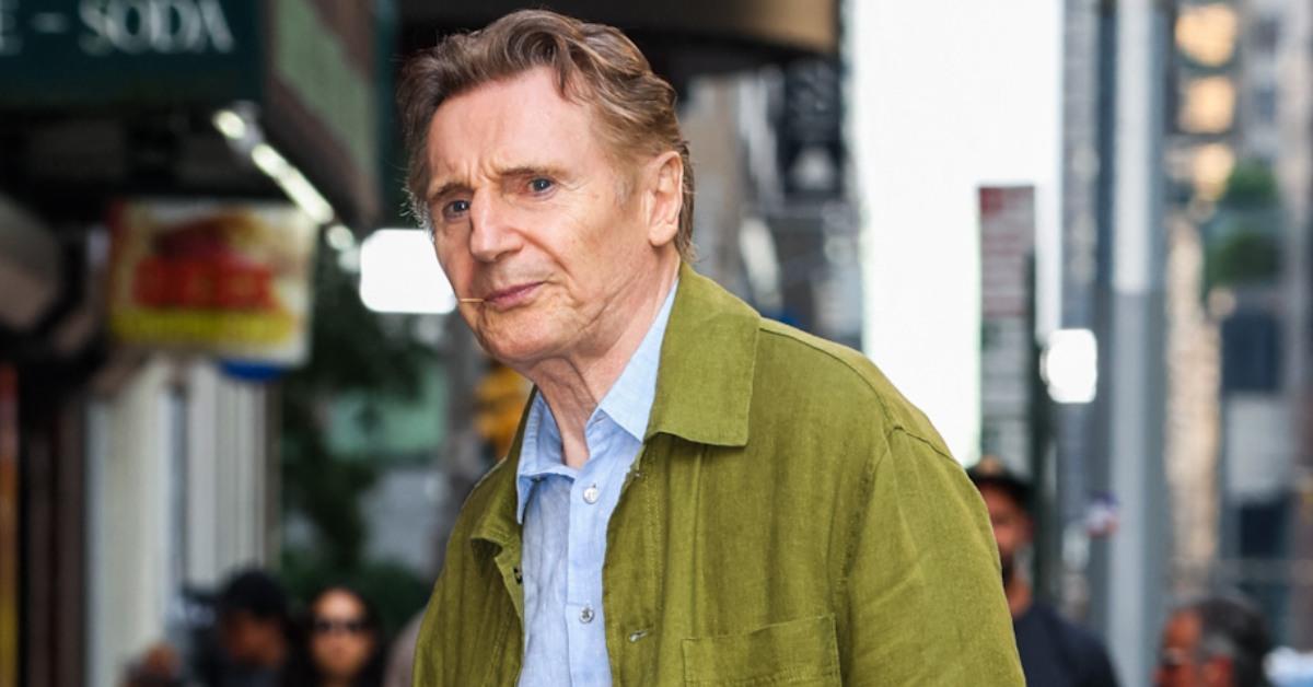 Liam Neeson wearing a green jacket casually walking around.