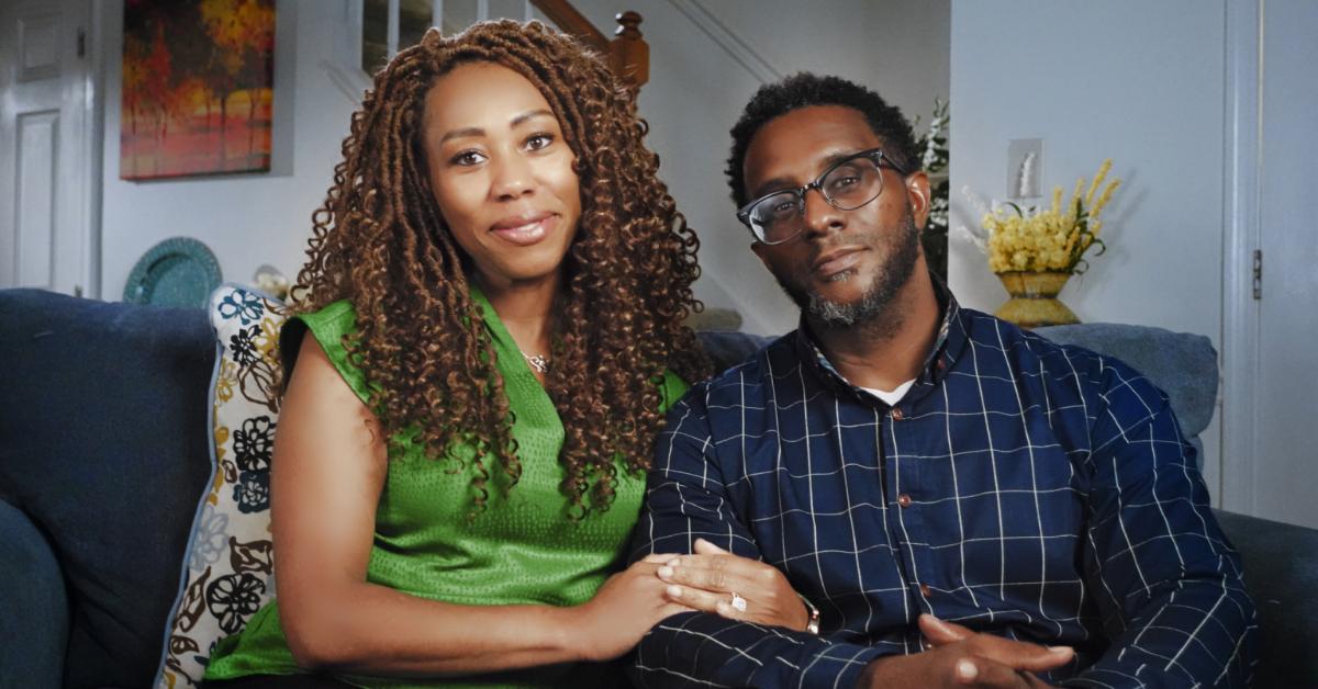 Nailah Salahuddin, dressed in a green shirt and black pants, sits on the couch with her arms intertwined with Naeem Salahuddin, who wears glasses and a blue and white dress shirt paired with light-colored pants.