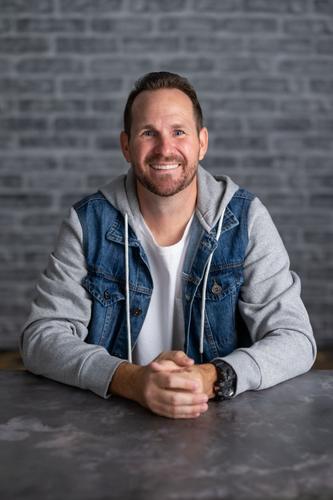 Sean is dressed in a white t-shirt and a denim jacket with a hood, smiling for his official portrait for Season 2 of 'The Mole.'