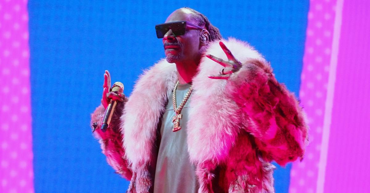 Snoop Dogg wears dark sunglasses massive pink fur coat with a green t-shirt underneath and throws up crip sign on stage while holding a microphone.
