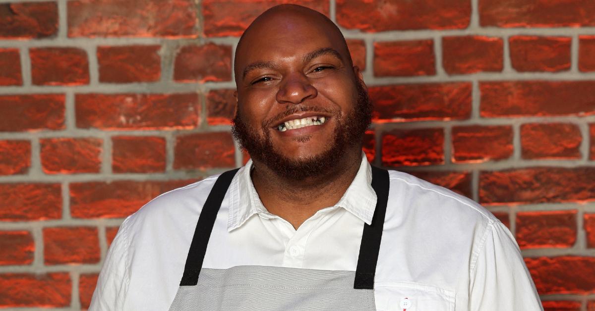 Ryan "Von" Smith from Season 3 of 'Next Level Chef' stands against a brick wall, dressed in a white button-up and a gray apron.