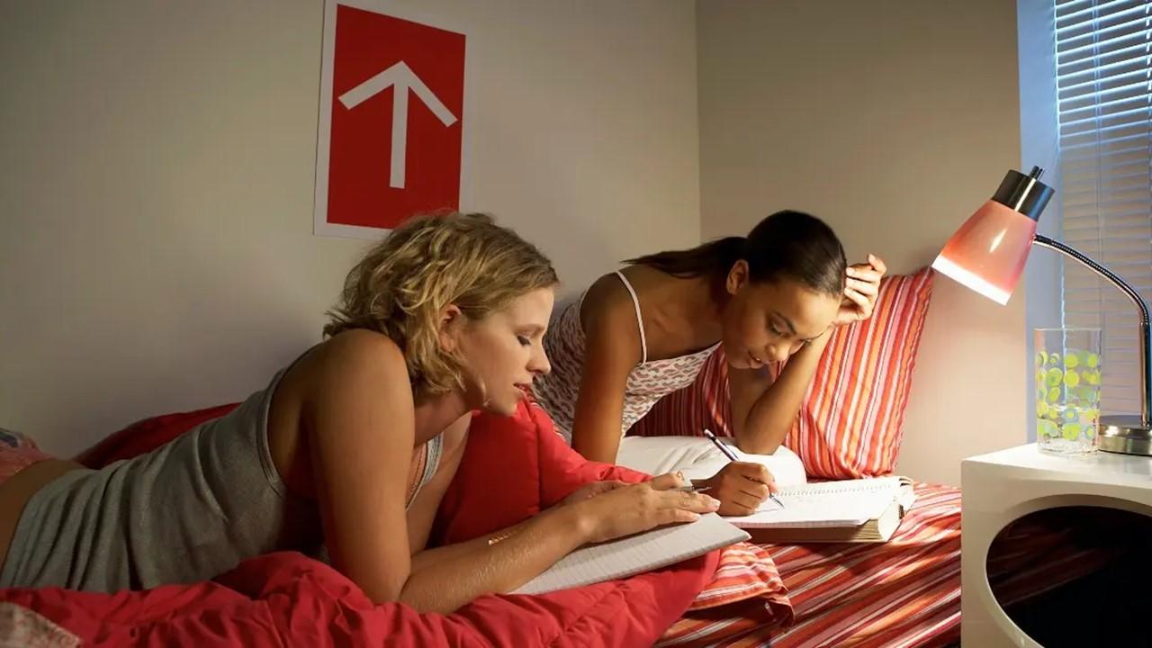 Two young women sitting on bed studying next to a lamp