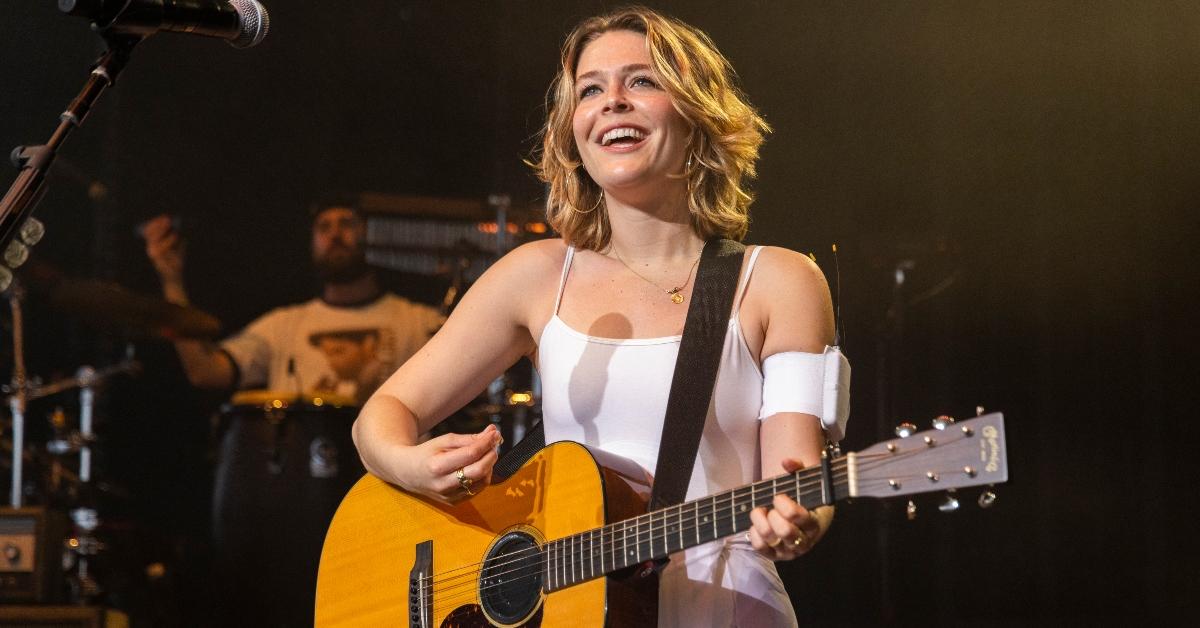 Maggie Rogers performs on stage in white dress holding her guitar - STERLING HEIGHTS, MICHIGAN - JUNE 09: Maggie Rogers performs at Michigan Lottery Amphitheatre on June 09, 2024 in Sterling Heights, Michigan. (Photo by Scott Legato/Getty Images)