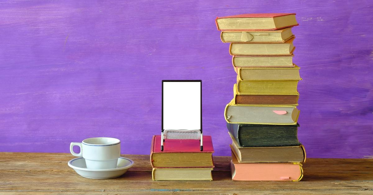 Stack of books alongside an e-reader in front of a purple wall background. 