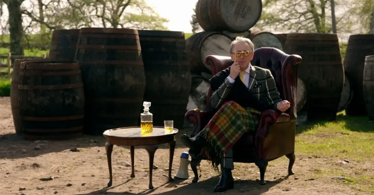 Alan Cumming sits on a chair in front of barrels outside on The Traitors