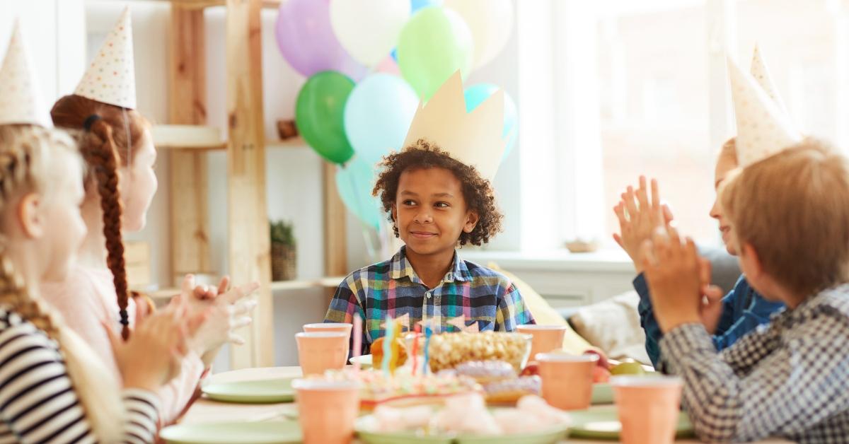 Little boy in plaid button-down shirt and birthday hat looks mischievous while sitting at a decorated table with his friends. 