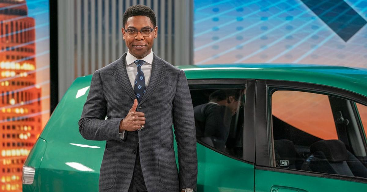 Michael Benjamin Washington as Cyrus in 'American Auto' wearing a grey suit, gives the thumbs up in front of a green van