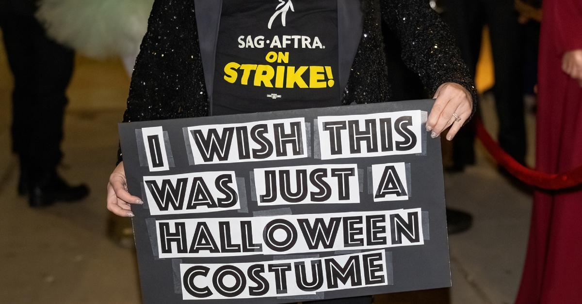 SAG-AFTRA member wears a "SAG-AFTRA on Strike" shirt while holding up a sign that reads, "I wish this was just a Halloween costume."