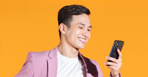 man with a pink blazer smiles and looks at his phone in front of an orange background