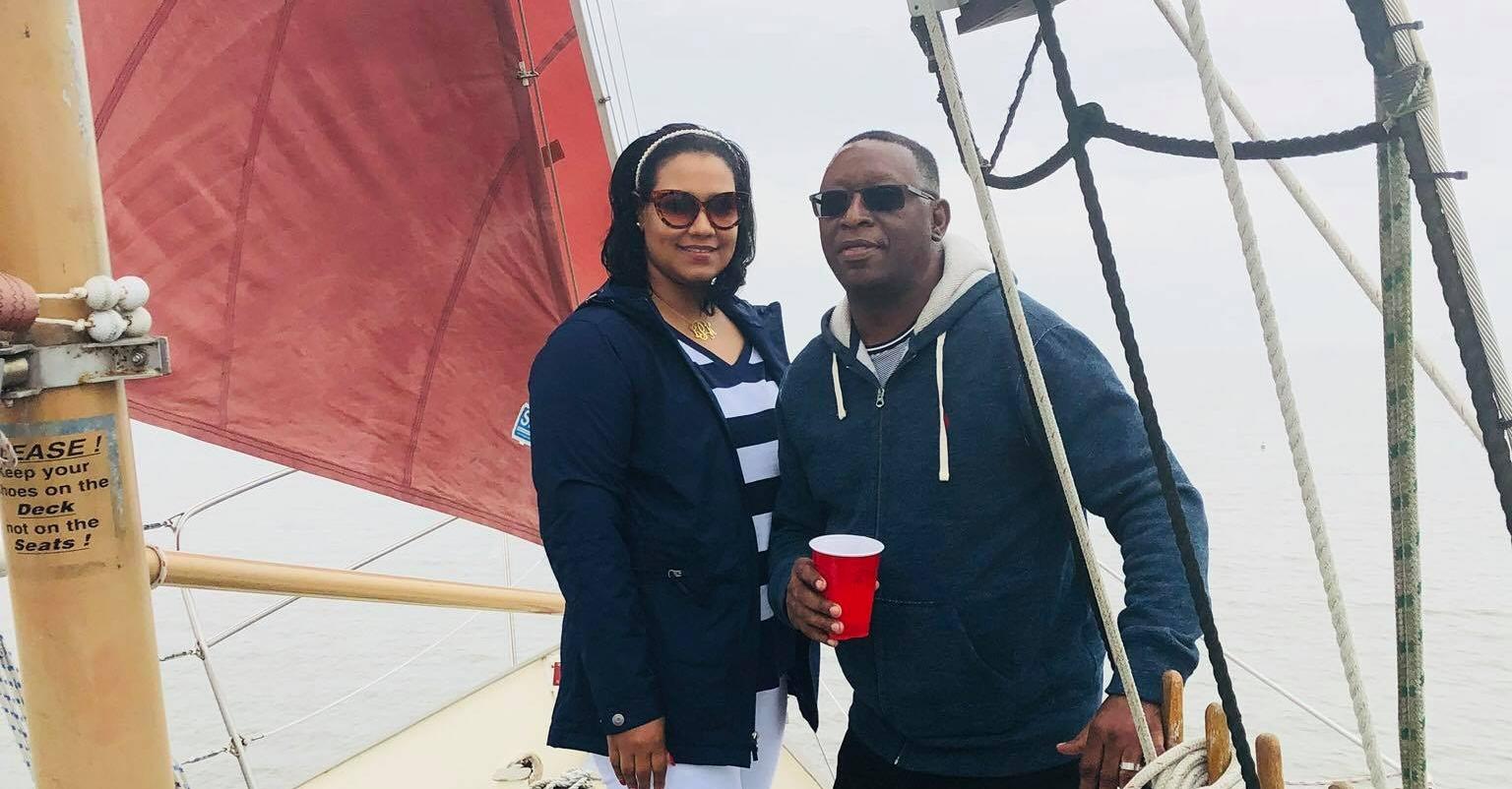 SheRa Seven and her husband pose on a sailboat wearing jackets.