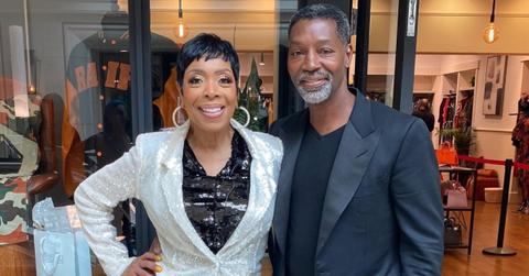 Shirley Strawberry wears a silver and black suit while Ernesto wears a Black blazer in front of storefront.