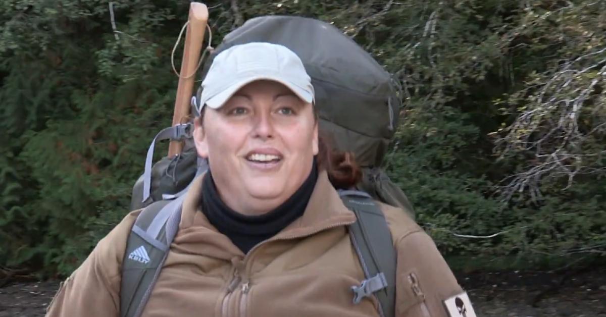 Tracy Wilson sports a cap, axe, backpack, and other survivalist gear in the outdoors.