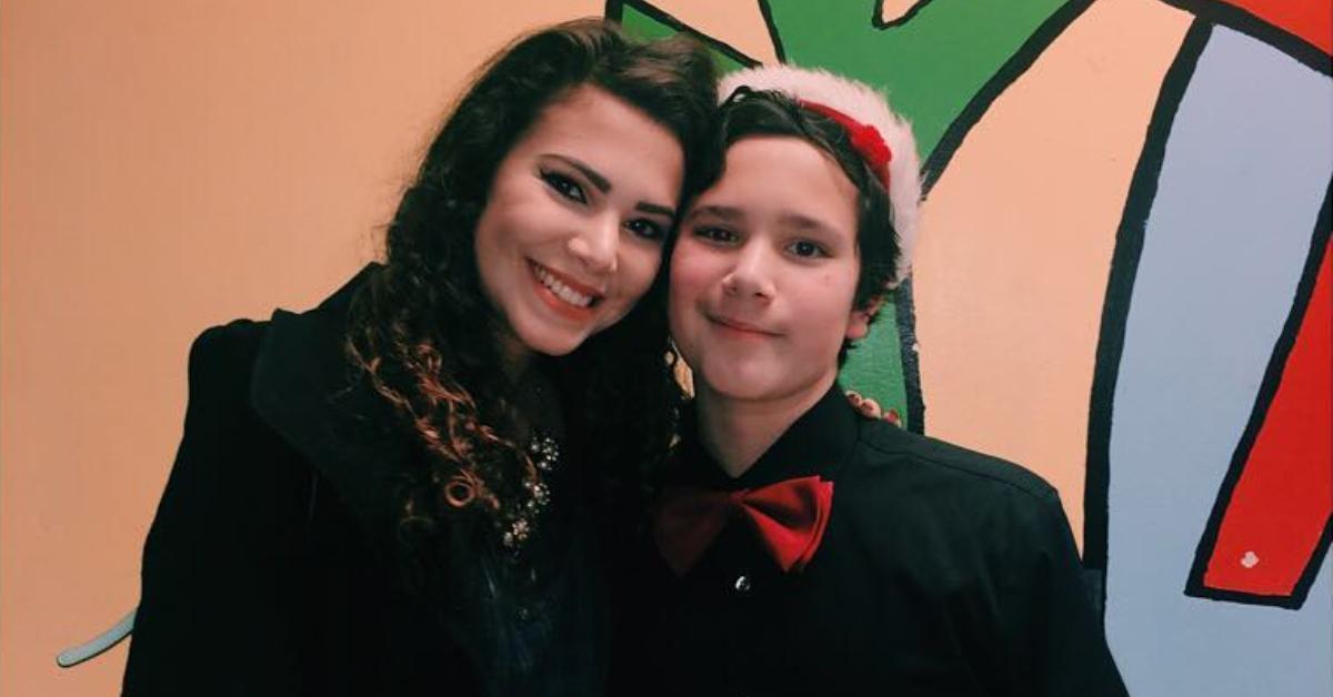 Amy Cortés grins and strikes a pose alongside her younger brother, David, who sports a black dress shirt paired with a vibrant red bowtie and a festive Santa hat.