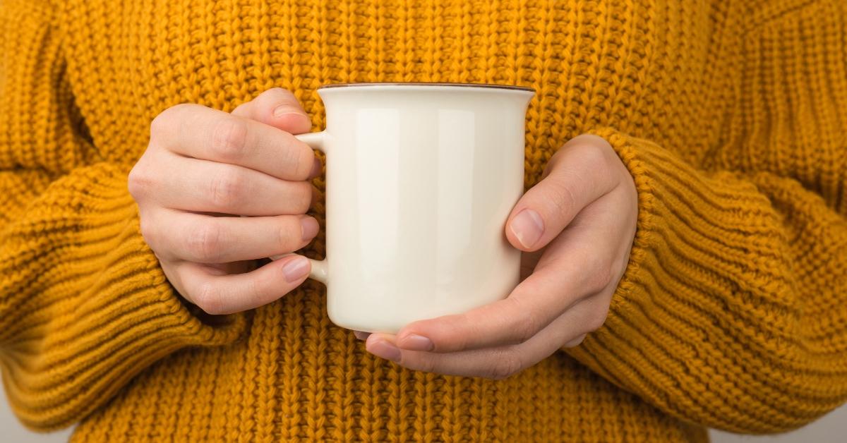Photo of young woman in yellow cozy sweater holding white mug of hot drink with empty space - stock photo