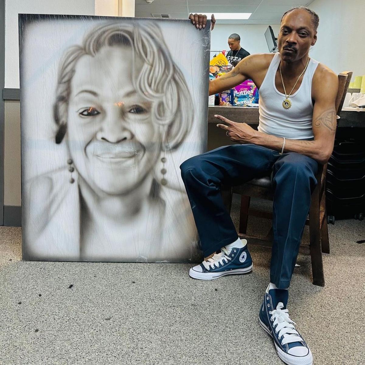 Snoop Dogg sits in a chair backstage holding a giant painting of his mom, Beverly Tate.