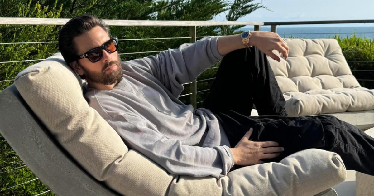 Scott, wearing a gray long-sleeve shirt and black pants, lounges outside on a chair.