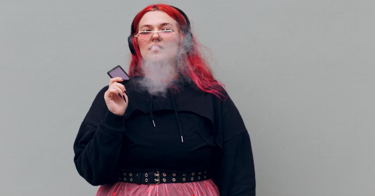 European plus size woman in headphones smoking vape electronic cigarette. Young red pink haired body positive girl - stock photo