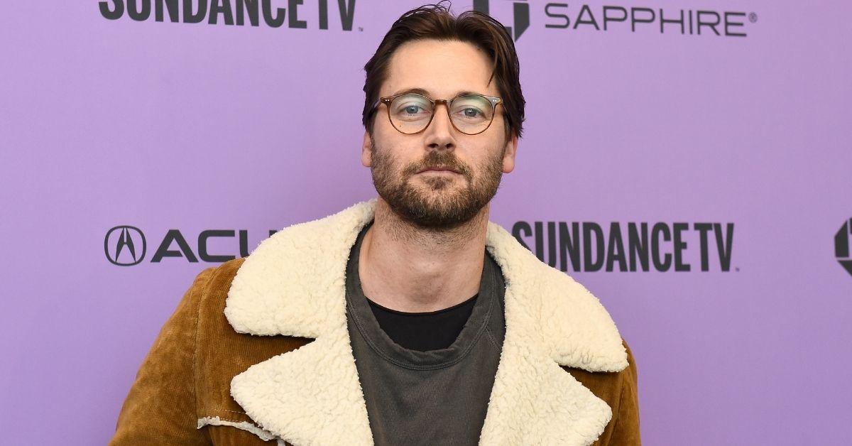 Ryan Eggold attending a red carpet event in glasses and a coat.