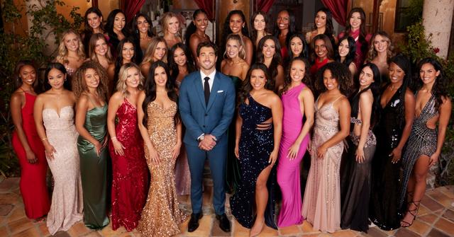In the Season 28 cast photo of 'The Bachelor,' Joey takes center stage, donning a sharp blue suit and black dress shoes, beaming with a smile. Surrounding him, the ladies grace the scene in an array of vibrant gowns, showcasing hues of red, pink, black, green, and gold.