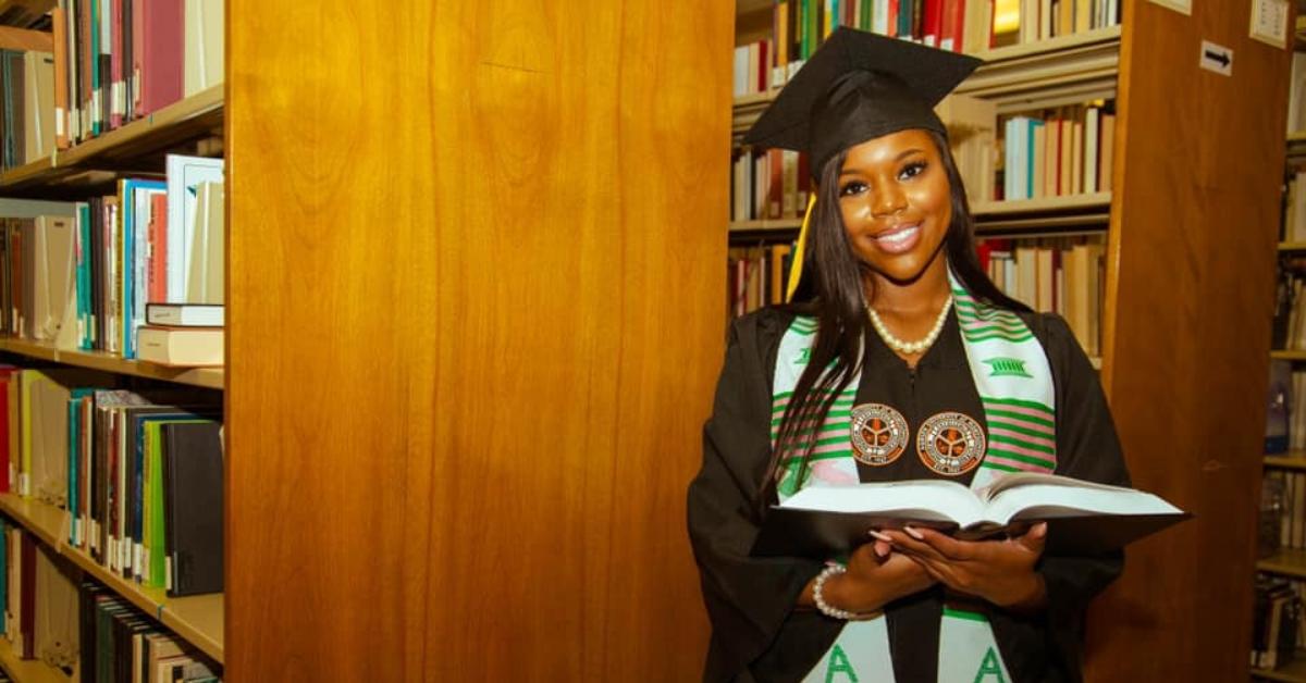 Carlee Russell wears a cap and gown and holds a book in library.