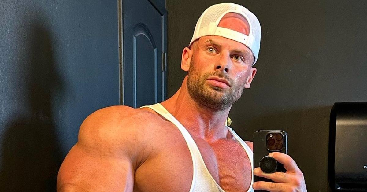 Joey Swole in a white tank top and hat poses in the mirror for a selfie with a serious face.