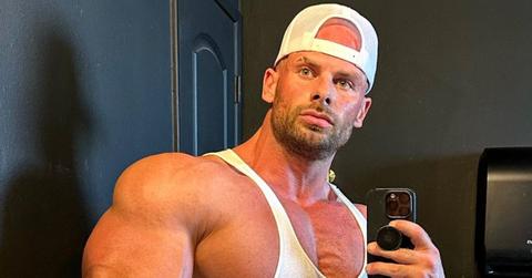 Joey Swole in a white tank top and hat poses in the mirror for a selfie with a serious face.
