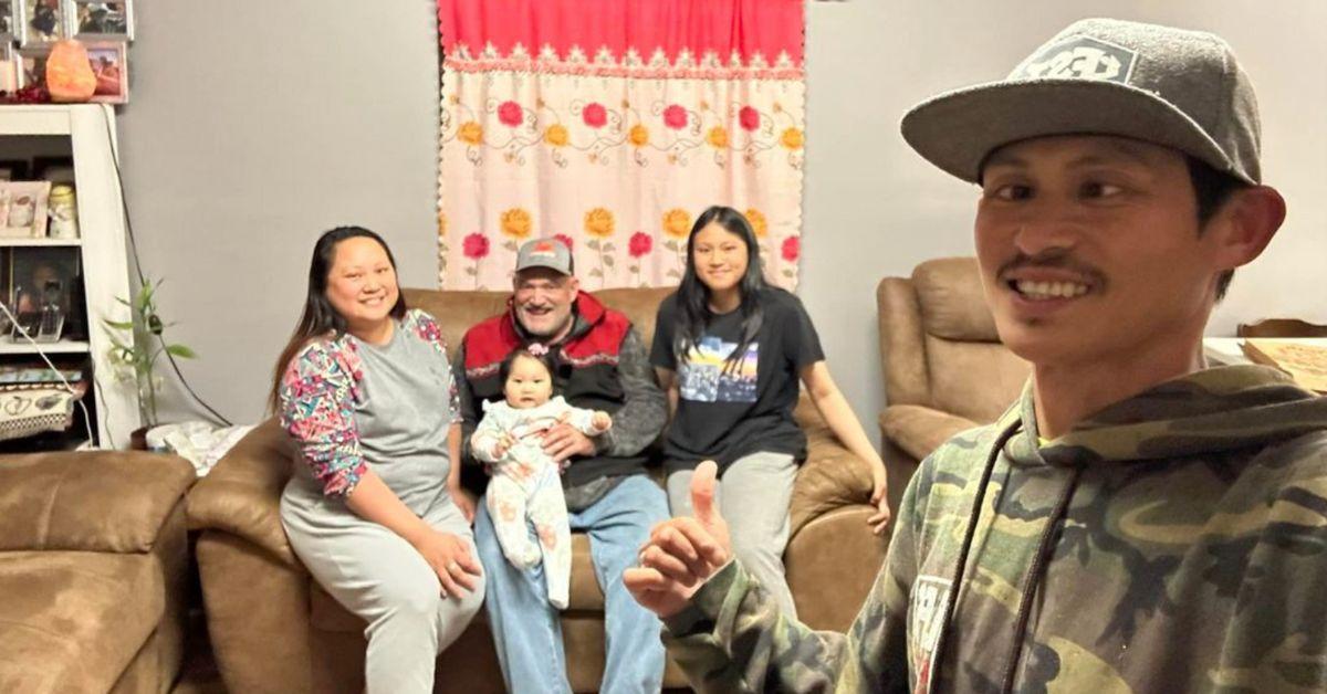 Oj (standing on the right) takes a selfie with his wife, Captain Keith who is holding Oj's youngest daughter, and oldest daughter all seated on the couch.