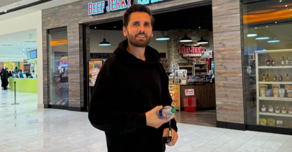 Scott, wearing an all-black sweatsuit, smiles and poses in front of a Beef Jerky store.