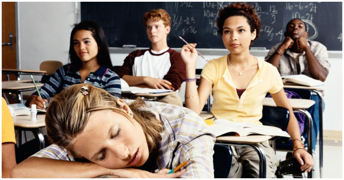 A student sleeping in a classroom as other students look at a board.