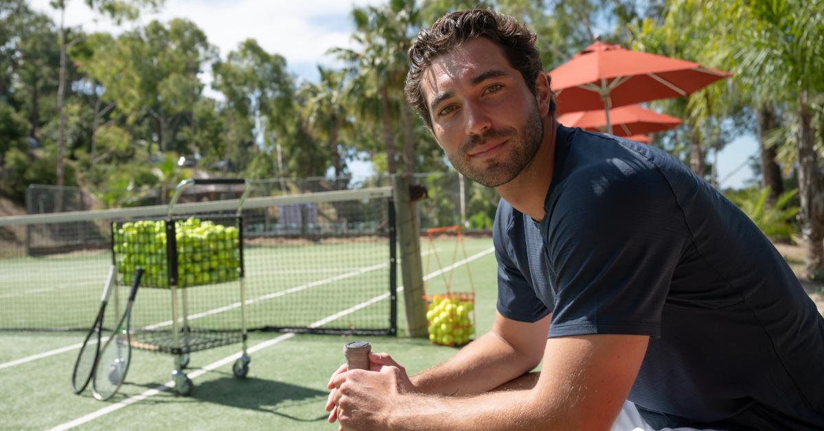 Joey Graziadei gazes into the camera, clad in white athletic shorts and a navy dri-fit shirt, seated on a bench overlooking a tennis court.