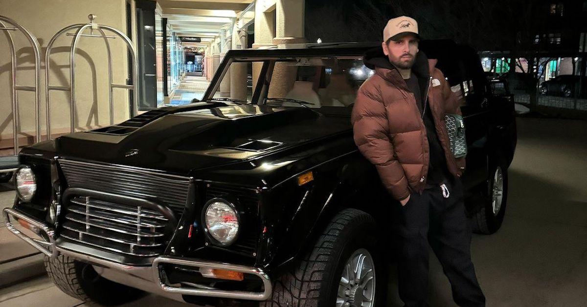 Scott Disick stands in front of his car wearing puffer jacket and hat