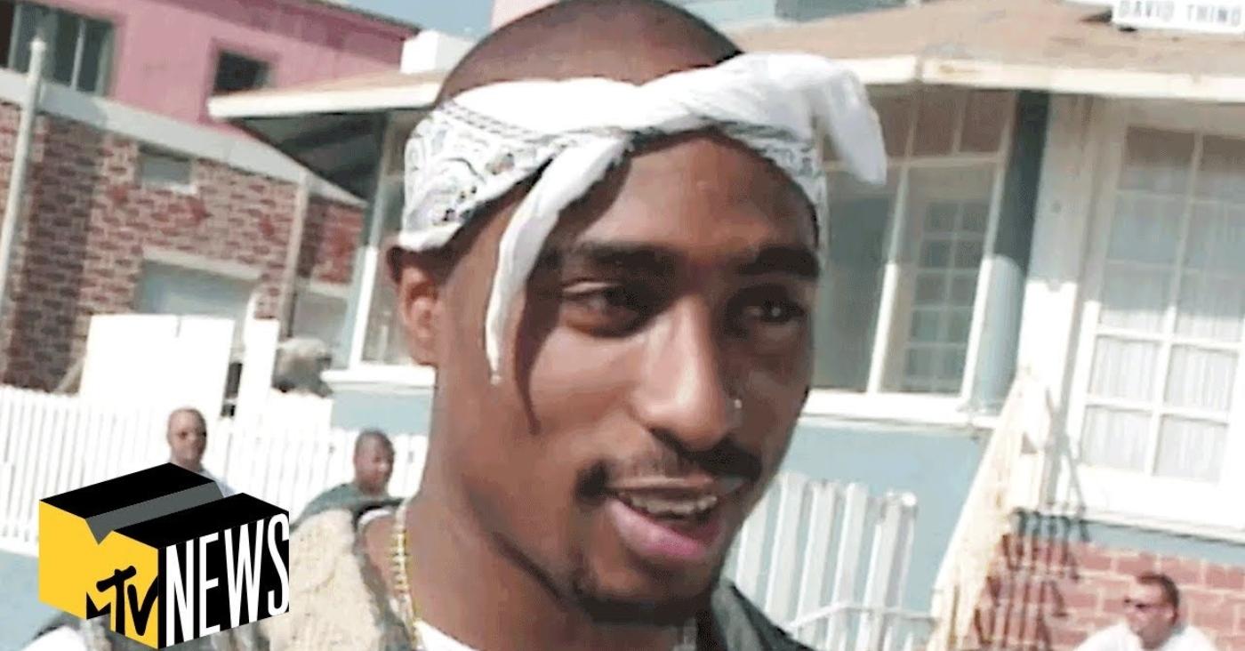 Tupac Shakur with a white bandana on in front of a house.