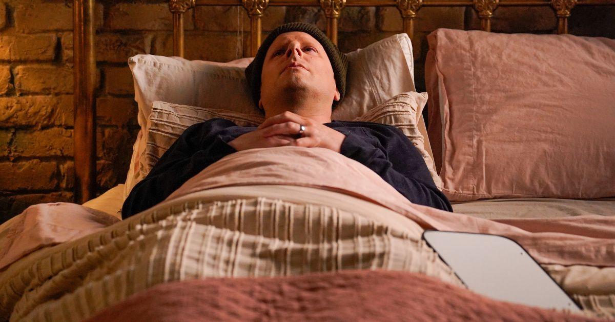 James Roday Rodriguez as Gary lying in bed with a beanie and long sleeved shirt.