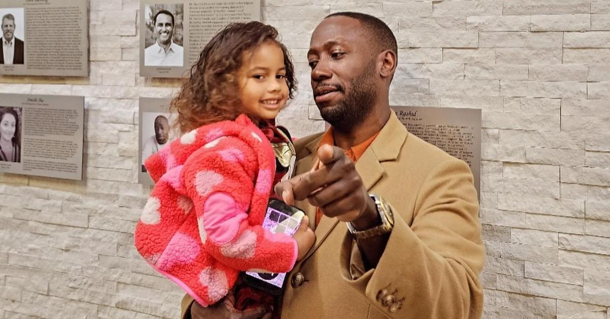 Lamorne Morris holding his daughter, Lily in his arms as he points to the camera.