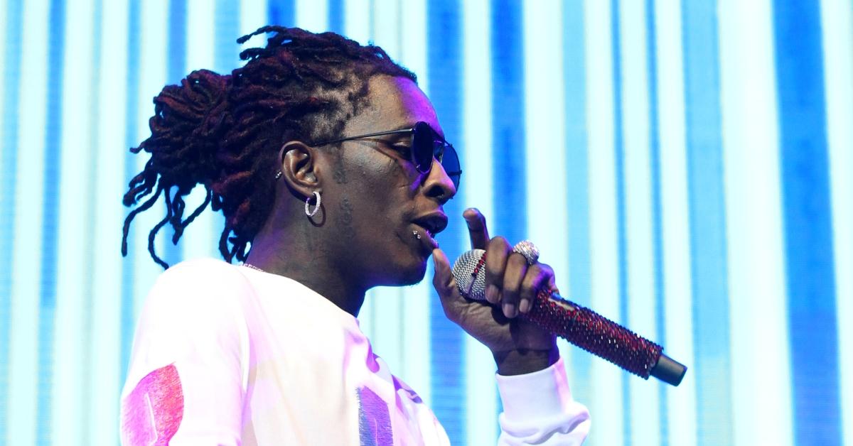 Young Thug rapping into a microphone in front of a neon blue background. 