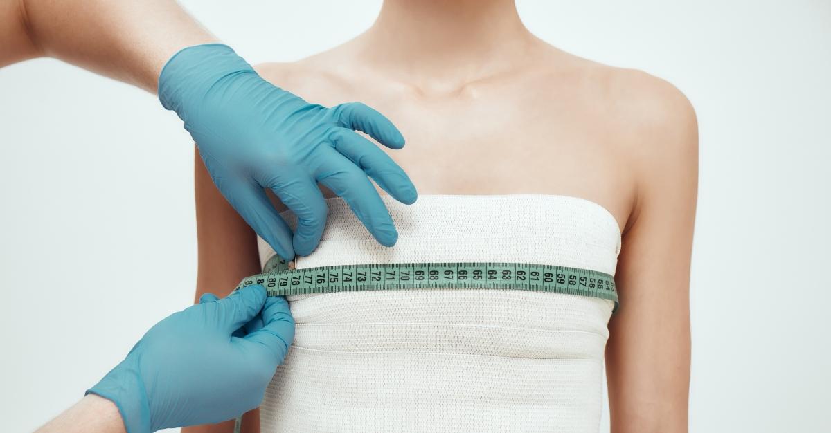 Breast augmentation operation. Cropped photo of woman waiting for plastic surgery while surgeons in blue medical gloves measuring her breast - stock photo