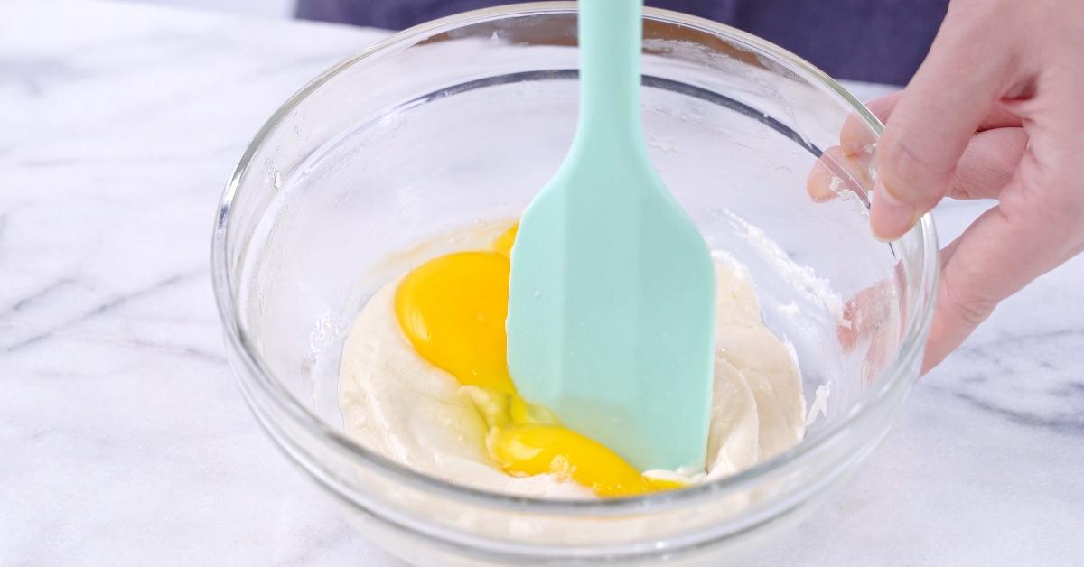 Mixing egg yolk into cake batter with green rubber spatula mixer tool stirring until smooth and blend well in a glass bowl. - stock photo
