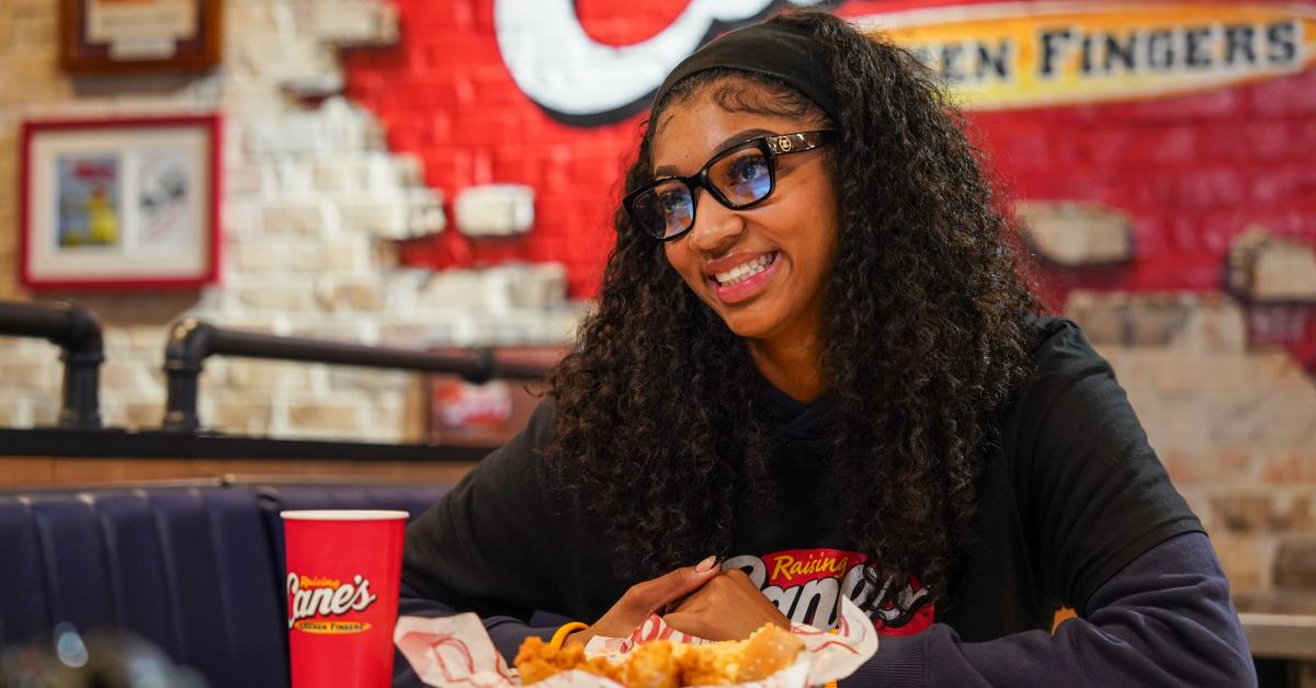 Angel Reese, wearing glasses and a Raising Cane's shirt over a sweater, smiles while looking away from the camera.