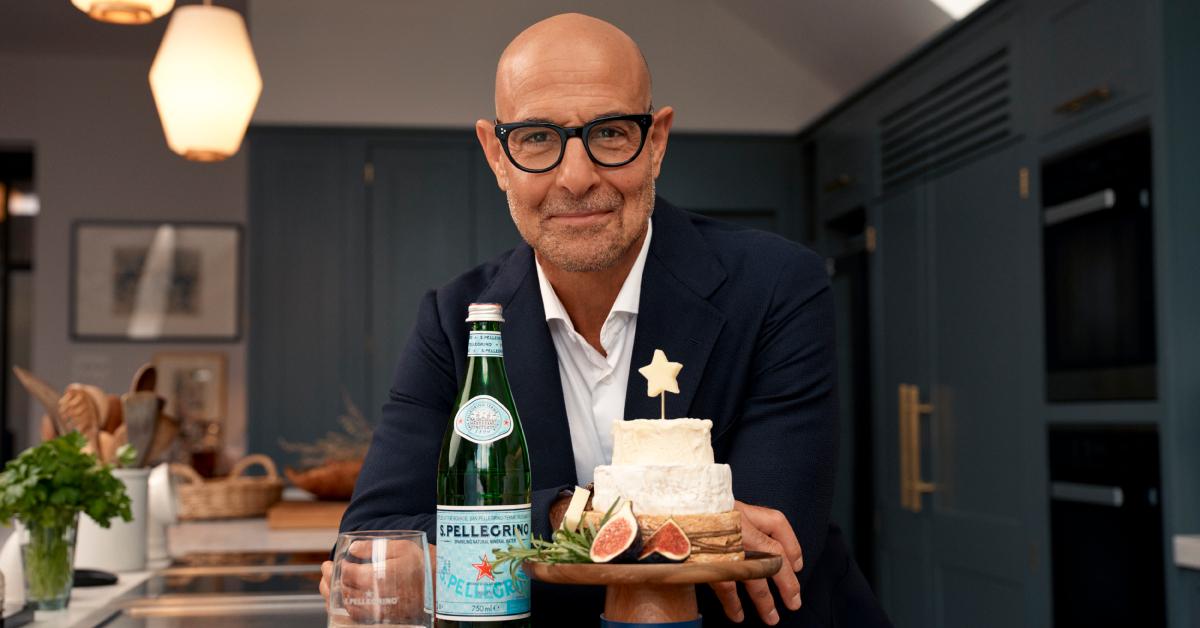 Stanley Tucci leaning on a table and posing with a bottle of S.Pellegrino and the "S.Pellegrino & Stanley Tucci's Holiday Cheese 'Cake'"