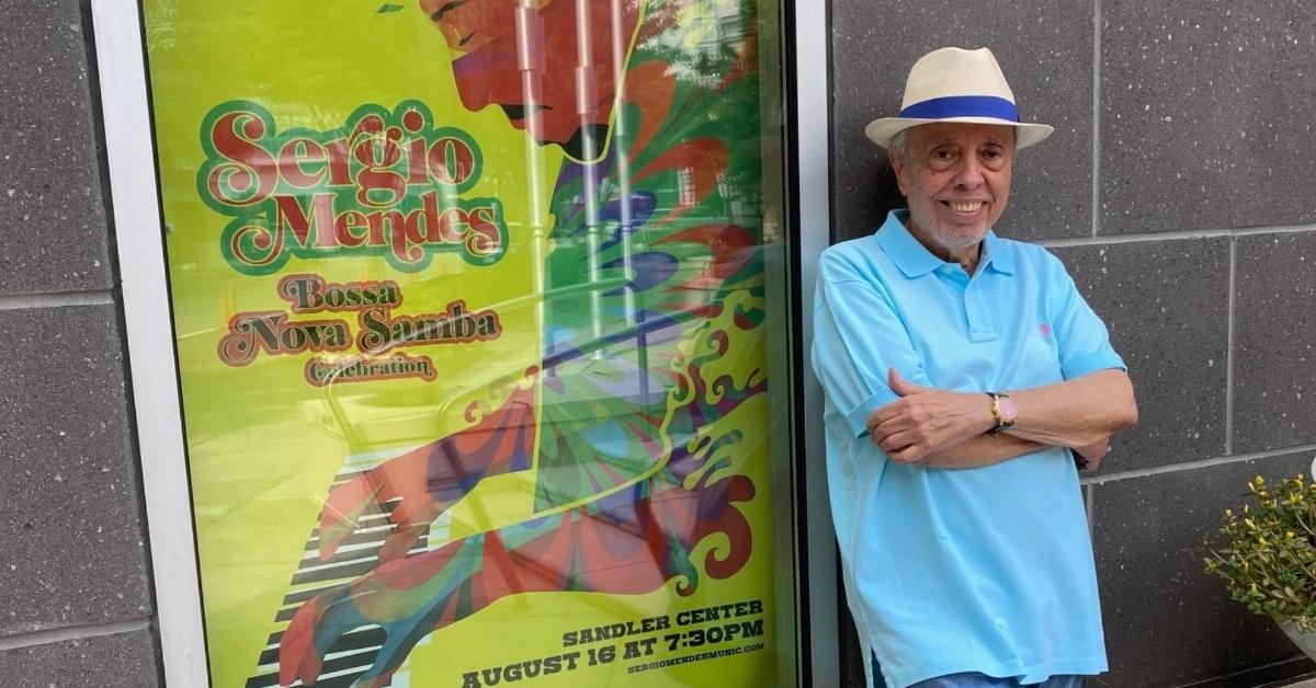 Sergio Mendes poses in blue shirt and hat in front of bossa nova samba concert music poster 2023