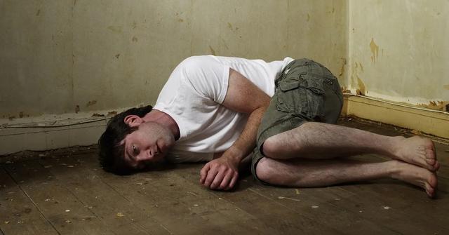 A man wearing a white shirt and green shorts curled up on the floor.