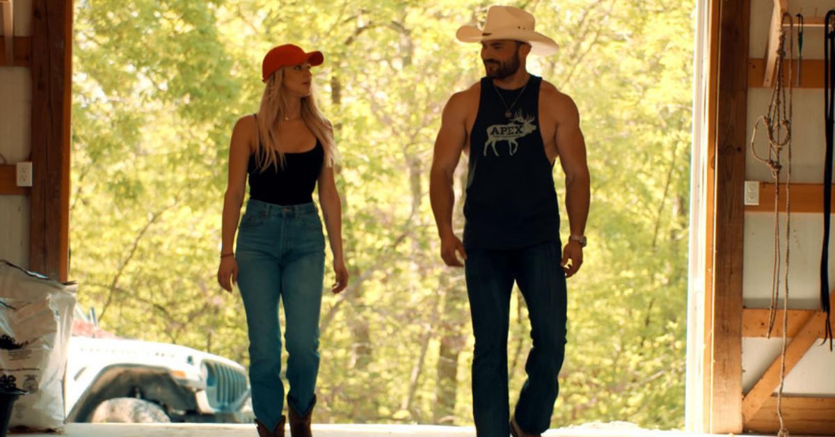 Calah Jackson wears a red baseball cap, a black tank top, and high-waisted jeans as she strolls next to Steven Jr. McBee, who dons a light-colored cowboy hat, a black muscle tank featuring an "Apex" logo, and black jeans.