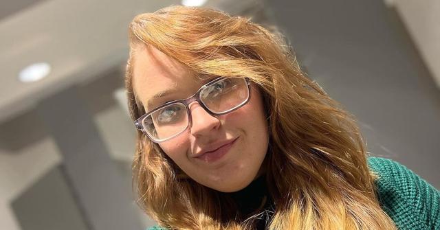 Anna wears glasses and a green sweater in a 2023 photo