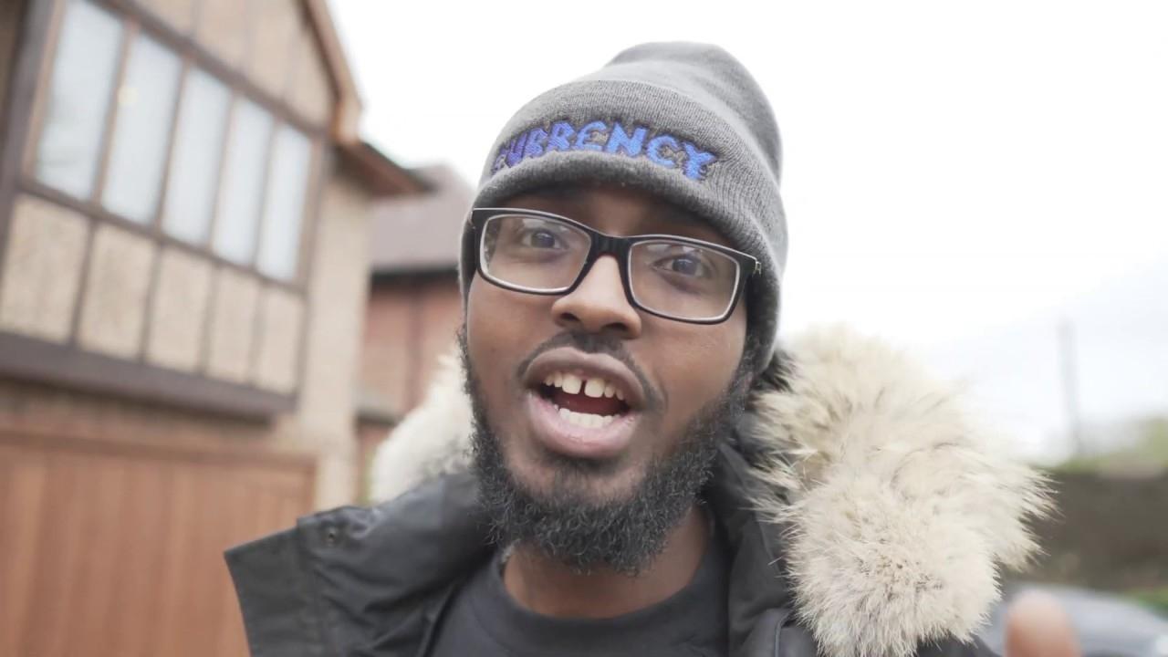 Darkest Man speaks in. a YouTube video wearing a gray beanie that says "CURRENCY", glasses, and a black puffer coat with fur on the hood.