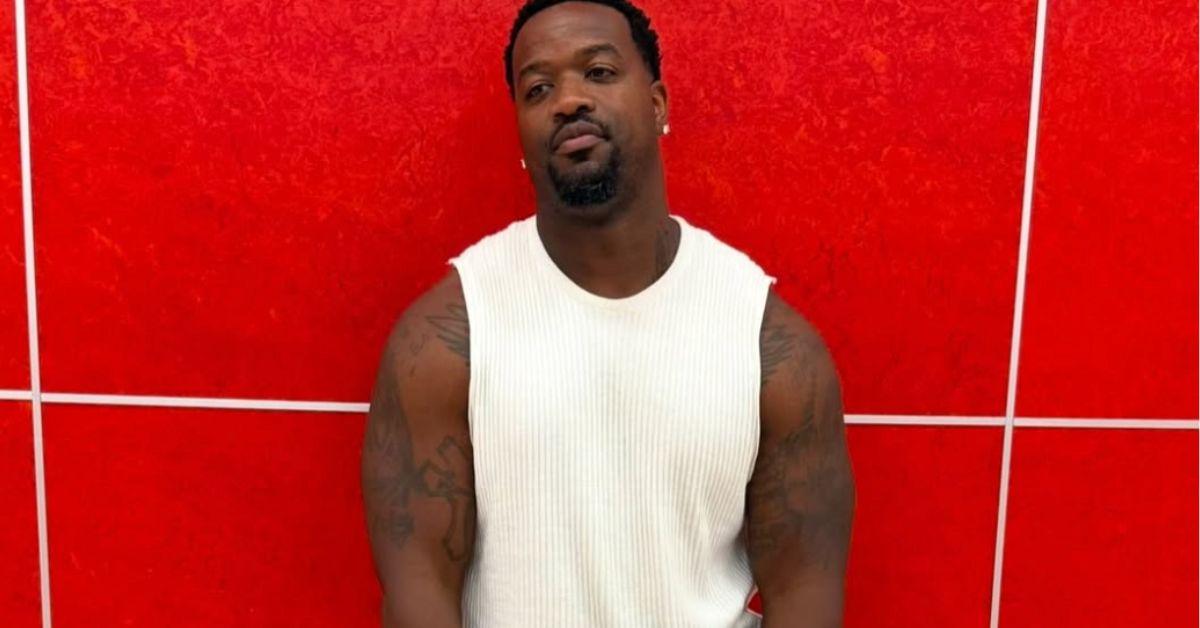 Jamar Champ standing against a red wall in a white tanktop. 