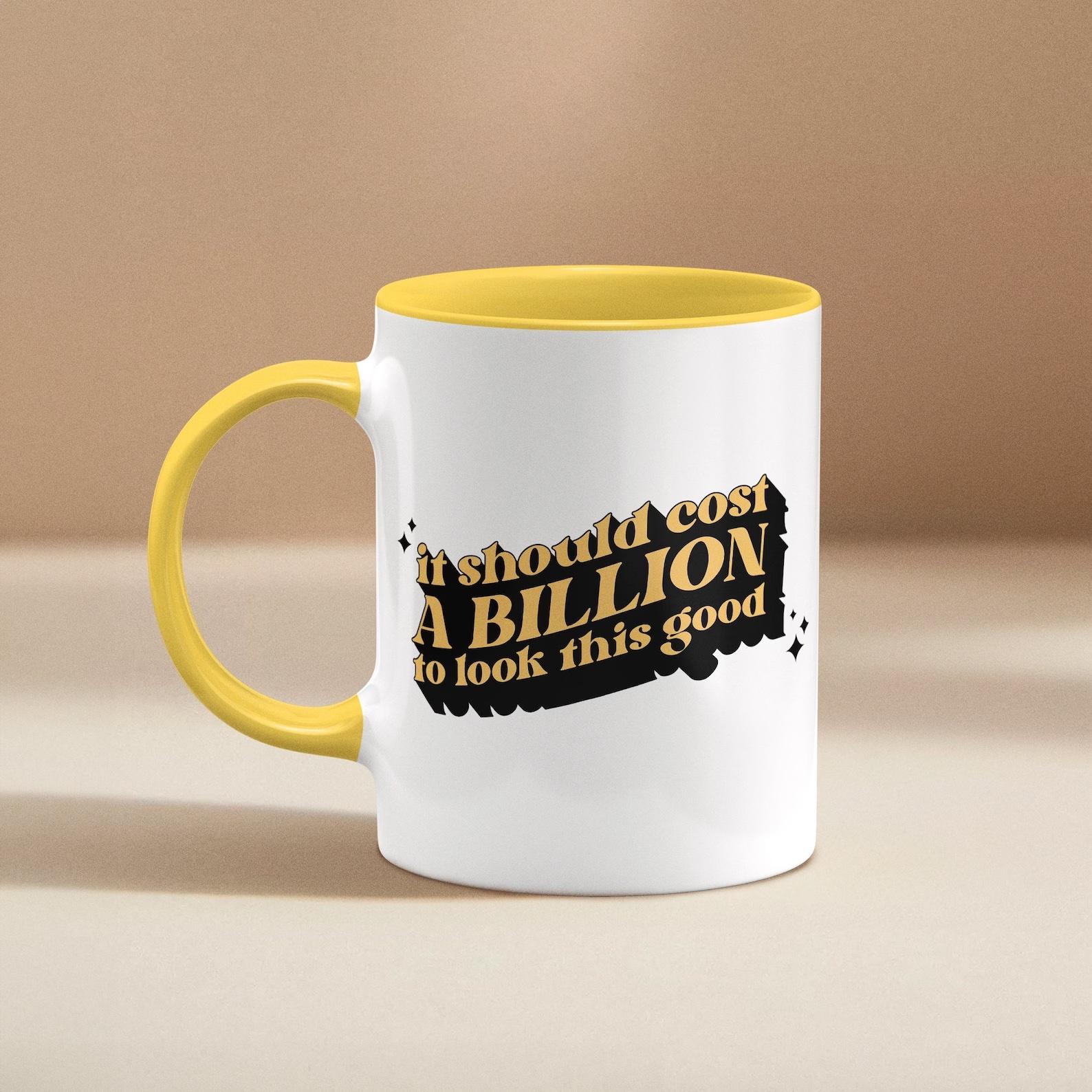 A while mug with a yellow handle and inside that reads "It should cost a billion to look this good"