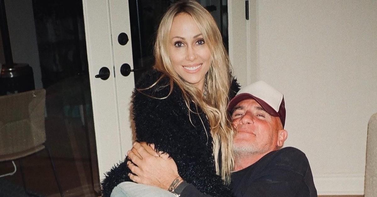 (L-R) Tish Cyrus in a black sweater, is embraced by Dominic Purcell in a black shirt and red and black baseball cap. 